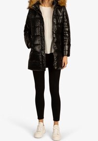 Doudoune noire avec une texture brillante, dotée d'une capuche en fausse fourrure, d'une fermeture éclair et d'un design matelassé. Associée à des leggings noirs et des baskets blanches.