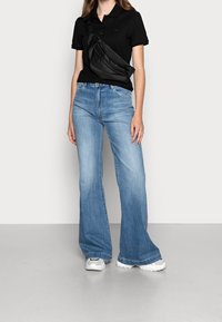 Polo shirt noir, jean large bleu clair et baskets blanches. Un sac bandoulière noir avec des zips est porté sur le torse.