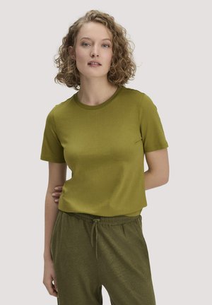 Frau mit lockigem blondem Haar, die ein olivgrünes T-Shirt und eine Hose mit Kordelzug trägt, steht mit einer Hand hinter dem Rücken.