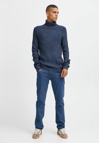 Mornarsko modra rebrasta turtleneck pulover, v kombinaciji z modrimi hlačami in sivo obutvijo. Pulover ima dolge rokave in prilegajoč silhueto.