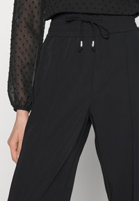 Calças pretas com uma cintura elástica e cordão, apresentando uma textura suave, bolsos laterais e um design de ajuste relaxado.