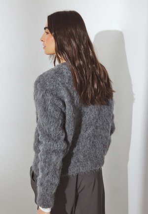 Suéter gris difuso con un acabado texturizado y esponjoso, con mangas largas, combinado con pantalones oscuros y holgados. Vista de perfil lateral.