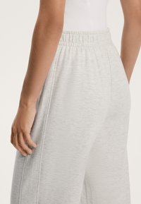 Svetlo sivi trenirke z teksturirano površino, elastičnim pasom in stranskimi žepi. Pogleduje se zadaj in del roke.