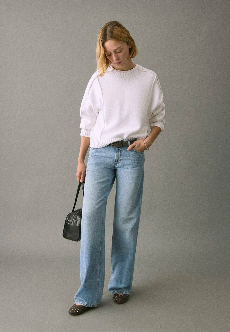 Weißer Strickpullover, hellblaue Jeans mit weitem Bein, schwarze Handtasche, braune Schuhe mit Nieten. Einfaches Design und lässige Farben.