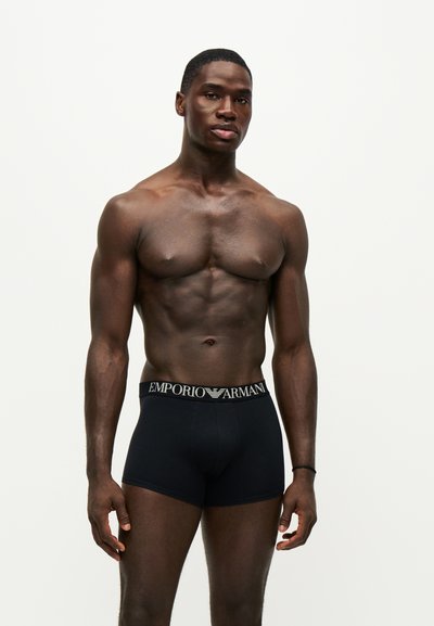 Homme musclé portant un boxer noir Emporio Armani, debout devant un fond clair uni, faisant face à l'objectif avec une expression neutre.