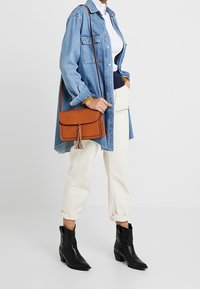 Brun skinnväska med axelrem och tofsdetalj. Bärs med en denimskjorta, vit topp och krämfärgade avsmalnande byxor. Svarta ankelboots.