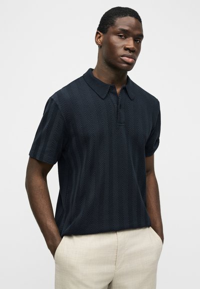 Άνδρας με κοντά μαλλιά που φοράει σκούρο πλεκτό κοντομάνικο πόλο και ανοιχτό μπεζ παντελόνι, στέκεται με τα χέρια στις τσέπες σε λευκό φόντο.