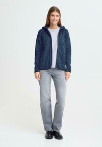 Tumši zilas krāsas kapuce ar rāvējslēdzēju, izgatavota no teksturēta auduma, kombinējoties ar gaiši pelēku kreklu un gaiši pelēkiem džinsiem. Tērpu papildina melna apavi.