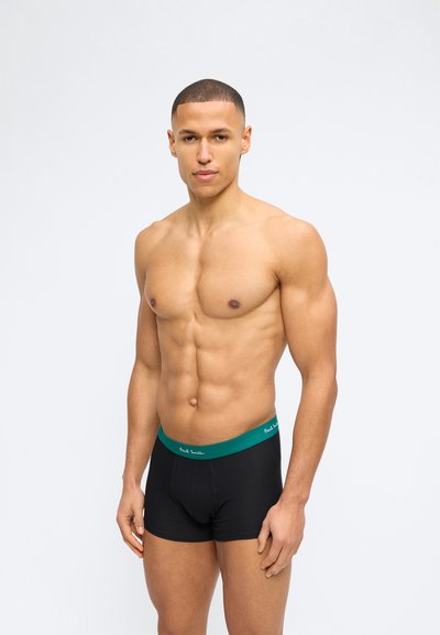 Μυώδης άντρας με κοντά μαλλιά που φοράει μαύρα εσώρουχα boxer με πράσινη ζώνη μέσης, στέκεται μπροστά σε ένα απλό λευκό φόντο.
