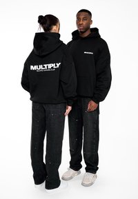 Felpe nere con la scritta "MULTIPLY" in bianco. Modello indossa un paio di pantaloni di jeans neri. Le felpe presentano tasche frontali e una vestibilità rilassata.