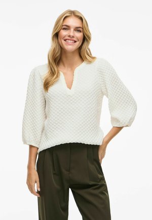 Pull en tricot blanc avec un décolleté en V, des manches trois-quarts et une coupe ample, associé à un pantalon taille haute vert foncé.