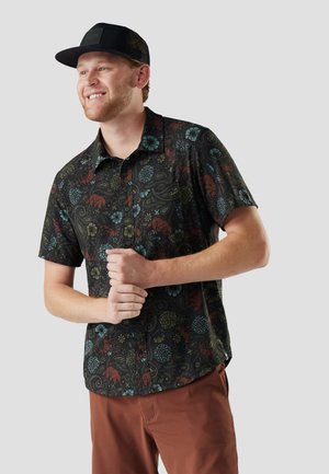 Ung mand smilende, iført sort kasket, mørk kortærmet skjorte med farverige dyre- og blomsterprint og rustfarvede bukser.