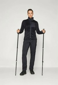 Casaco de fleece em azul escuro com fecho de zíper na frente, combinado com calças escuras e bastões de trekking. Textura suave, ajuste atlético e design funcional.