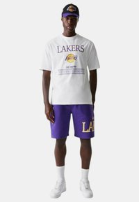 T-shirt bianca con grafica "LAKERS" e pantaloni corti viola con "LAKERS" in giallo. Abbinati a un berretto nero con accenti del logo. Sneakers bianche.