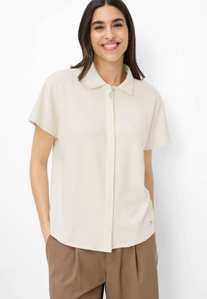 Lächelnde Frau mit langen dunklen Haaren, die ein cremefarbenes kurzärmeliges Hemd mit Knöpfen und braune, plissierte Hose trägt, mit den Händen in den Taschen.