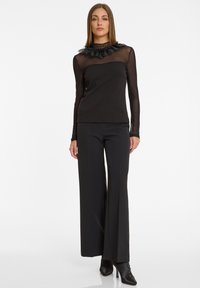 Schwarzes langärmliges Oberteil mit einem oberen Teil aus Netz und Rüschenkragen, kombiniert mit hochtaillierten, weiten schwarzen Hosen und Stiefeletten.