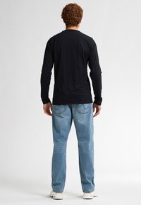 Schwarzes Langarmshirt kombiniert mit hellblauen Jeans. Das Shirt hat einen schlichten Rücken; die Jeans verfügen über Gesäßtaschen und eine lässige Passform. Weiße Sneakers sind sichtbar.