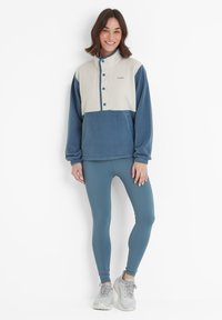 Woman standing and smiling wearing a blue and white fleece pullover, blue leggings, and white sneakers against a white background.