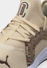 Μπεζ αθλητικό παπούτσι Puma με υφασμάτινη υφή, κορδόνια στον ίδιο χρωματισμό, καφέ λογότυπο στη γλώσσα και στην μύτη, και διάφανη μαρμάρινη πλευρική στήριξη.