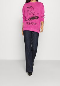 Lyserød sweatshirt med et Snoopy-tryk, parret med mørkeblå vidde jeans og sorte ankelstøvler. Sweatshirten har ribbede manchetter og rund halsudskæring.