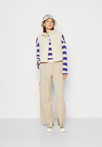 Beige fleece vest over een paars-witte gestreepte blouse met lange mouwen, gecombineerd met beige wijde broek en groene sneakers. Gelaagde bucket hat.