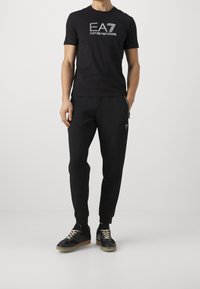 Svart kortärmad t-shirt med EA7 Emporio Armani-logotyp, parat med svarta träningsbyxor och svarta sneakers med beige snören och gummisulor.