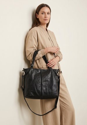 Vrouw in beige outfit leunt tegen de muur, met een grote zwarte leren tas met handvatten en schouderriem.