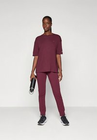 Bordový set športového oblečenia pozostávajúci z voľného krátkeho trička s krátkym rukávom a úzkych nohavíc, doplnený čiernymi športovými topánkami a fľašou na vodu.