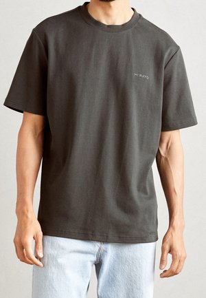 Mann in einem schlichten dunkelgrauen T-Shirt mit kleinem "Morato"-Logo auf der Brust und hellblauen Jeans, steht vor einem neutralen Hintergrund.