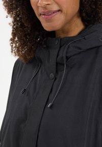 Giacca nera con cappuccio e coulisse regolabili. Presenta bottoni a pressione e una texture liscia e opaca. Primo piano dell'area del colletto.