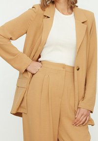 Blazer beige con una tela ligeramente texturizada, solapas con muesca y cierre de un botón, combinado con una blusa blanca simple y pantalones a juego.