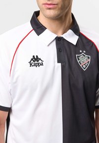 Homme portant un polo Kappa noir et blanc à col avec un liseré rouge et un badge Kappa Authentic sur la poitrine.