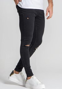 Jeans ajustados negros con detalles desgastados y aberturas deshilachadas en las rodillas, combinados con zapatillas blancas con acentos dorados.