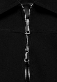 Veste noire zippée avec un col structuré, dotée d'une fermeture éclair métallique à deux curseurs et d'une texture de tissu lisse.