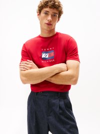 Tricou din bumbac roșu cu grafică "TOMMY HILFIGER" și "1985" în culorile albastru și alb. Modelul poartă pantaloni plisati albastru închis.