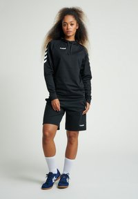 Svart sportig hoodie med vitt logotyp och tre vita chevron-ränder på ärmarna, ihopkopplad med svarta shorts. Blå sneakers och vita strumpor syns.
