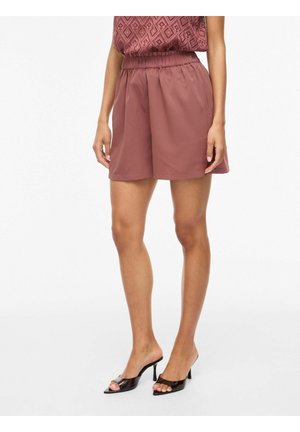 Kobieta ubrana w wysokiej talii mauve szorty z elastycznym pasem, zestawione z koronkową bluzką i czarnymi, otwartymi na palce, na obcasie mule.