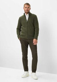 Donkergroene gebreide cardigan met hoge kraag, geribde manchetten en zoom, gecombineerd met bruine broek en witte sneakers. Eenvoudig, gestructureerd ontwerp.