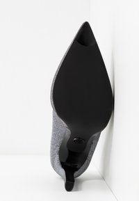 Chaussure à talon haut à bout pointu, semelle noire et dessus en tissu gris texturé, photographiée par le dessous sur fond blanc.