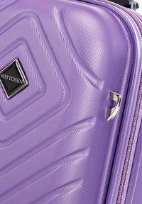 Maleta rígida morada con una superficie texturizada, que presenta una placa con logotipo negra y un acento plateado, diseñada con patrones en forma de ondas.