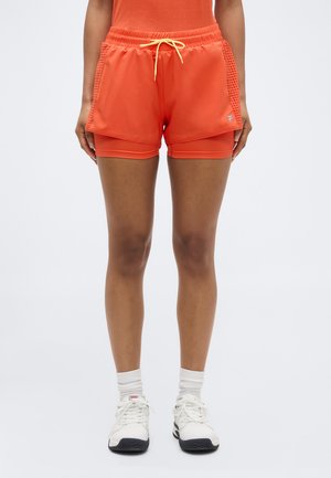 Persoon draagt feloranje gestapelde sportshorts met gele trekkoord, witte sokken tot aan de enkels en witte sneakers, staand tegen een effen achtergrond.