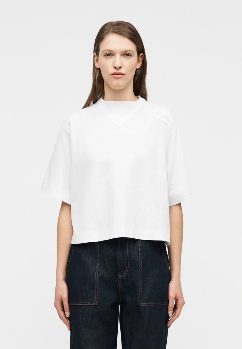 Γυναίκα που στέκεται μπροστά σε λευκό φόντο, φορ ώντας ένα άνετο λευκό κοντομάνικο τοπ και σκούρο ψηλόμεσο παντελόνι με εμφανείς ραφές.