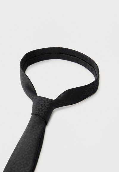 Cravate en soie texturée noire avec un nœud classique, posée à plat sur un fond gris clair.