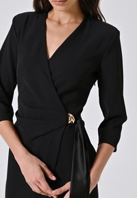 Frau, die ein schwarzes Wickelkleid mit drei Viertelärmeln und einem goldenen Verschluss in der Taille trägt, mit sanft gefalteten Händen vor sich.