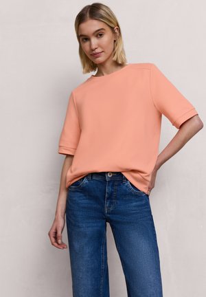 Junge Frau mit kurzen blonden Haaren, die ein lachsfarbenes Kurzarmoberteil und blaue Jeans trägt, steht vor einer schlichten hellen Wand.