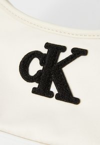 Étiquette logo Calvin Klein en noir brodée sur un fond de tissu crème clair, avec des détails de couture visibles autour des bords.