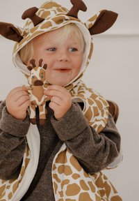 Bērns valkā džemperi ar žirafu rakstu un brūnām detaļām, turot rokās mīkstu žirafes rotaļlietu. Apģērbam ir mīksta tekstūra un rotaļīgs dizains.