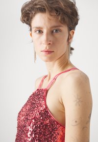 Särava halter-top roosa tooni, millel on õhukesed rihmad ja vormistatud disain. Silmapaistev läikiv tekstuur koos keeruka detailiga.