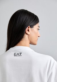Bijela pamučna majica s crnim EA7 Emporio Armani logotipom na stražnjoj strani izreza. Jednostavan dizajn s okruglim izrezom i kratkim rukavima.