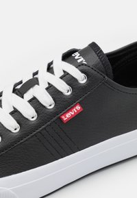 Sneaker in pelle nera con lacci bianchi e suola in gomma. Presenta un'etichetta rossa del logo Levi's e occhielli argentati. Superficie testurizzata con dettagli di cucitura.